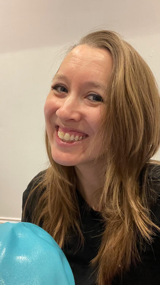 A portrait photo of Louise in a black T-shirt, smiling at the camera and holding a turquoise pilates ball